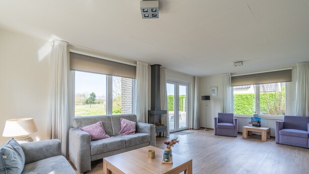 Een ruime woonkamer met een grijze bank, een houten salontafel en twee fauteuils. Er is een open haard, een groot raam met gordijnen en uitzicht op groen.