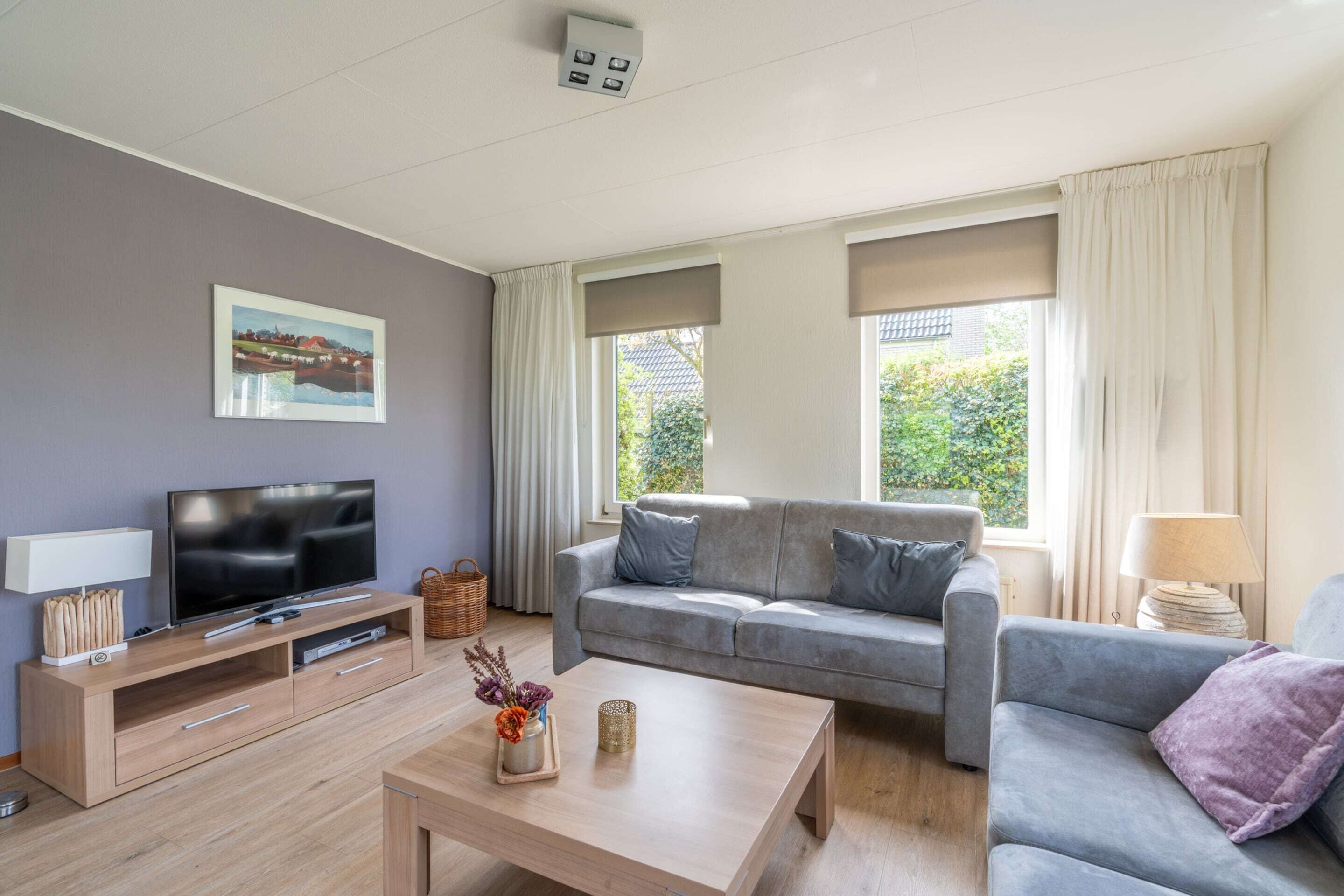 Una sala moderna con un sofá gris, un televisor de pantalla plana sobre un mueble de madera, una mesa de centro y ventanas amplias con cortinas.