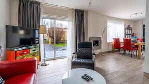 A modern living room with a flat-screen TV, a red sofa, a black leather chair, a dining area with red chairs, and a fireplace. - Cozy and Comfortable 4-person Bungalow (De Koog)
