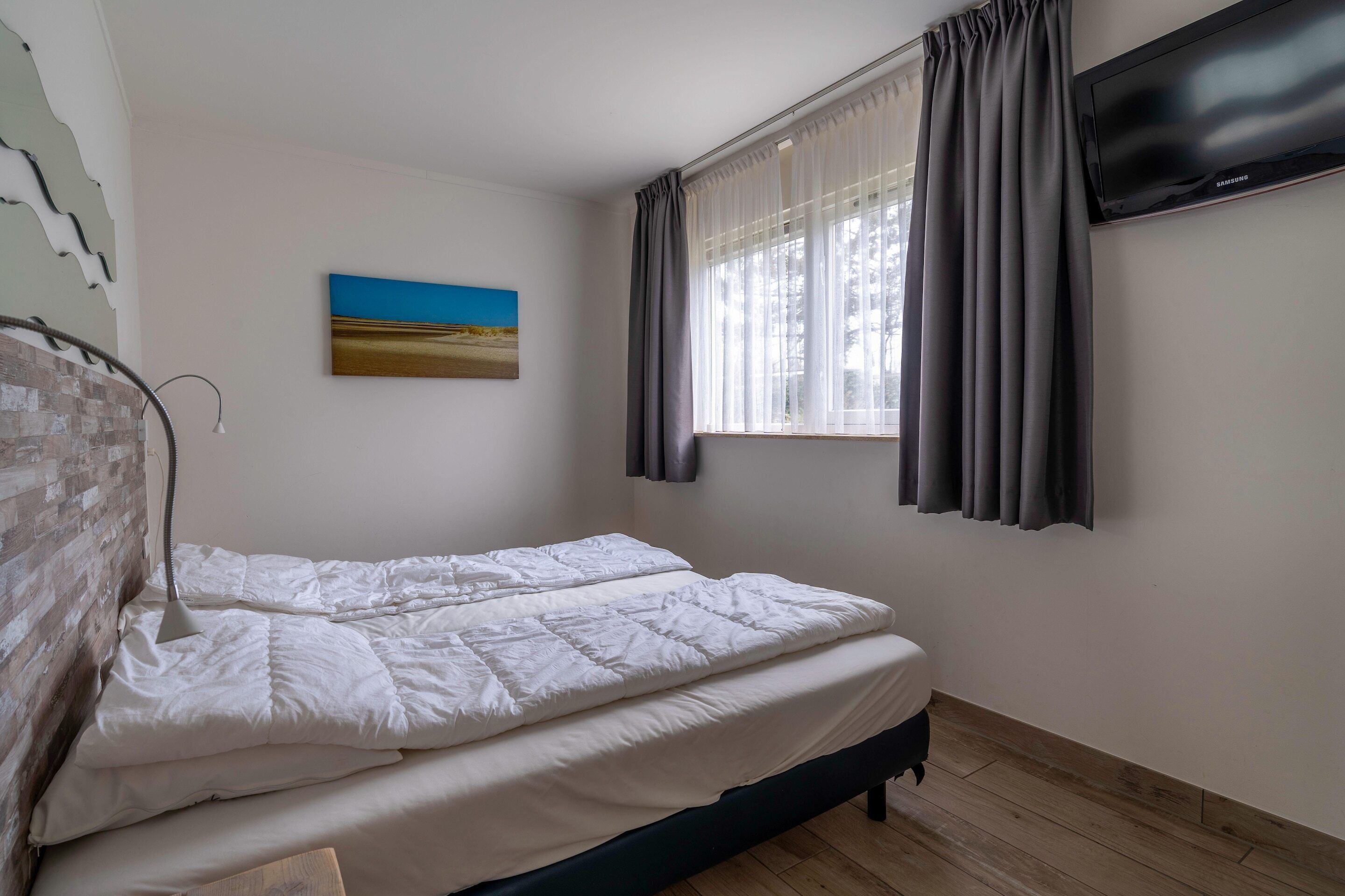 Un dormitorio con una cama, una ventana con cortinas, un televisor de pantalla plana y un cuadro en la pared.