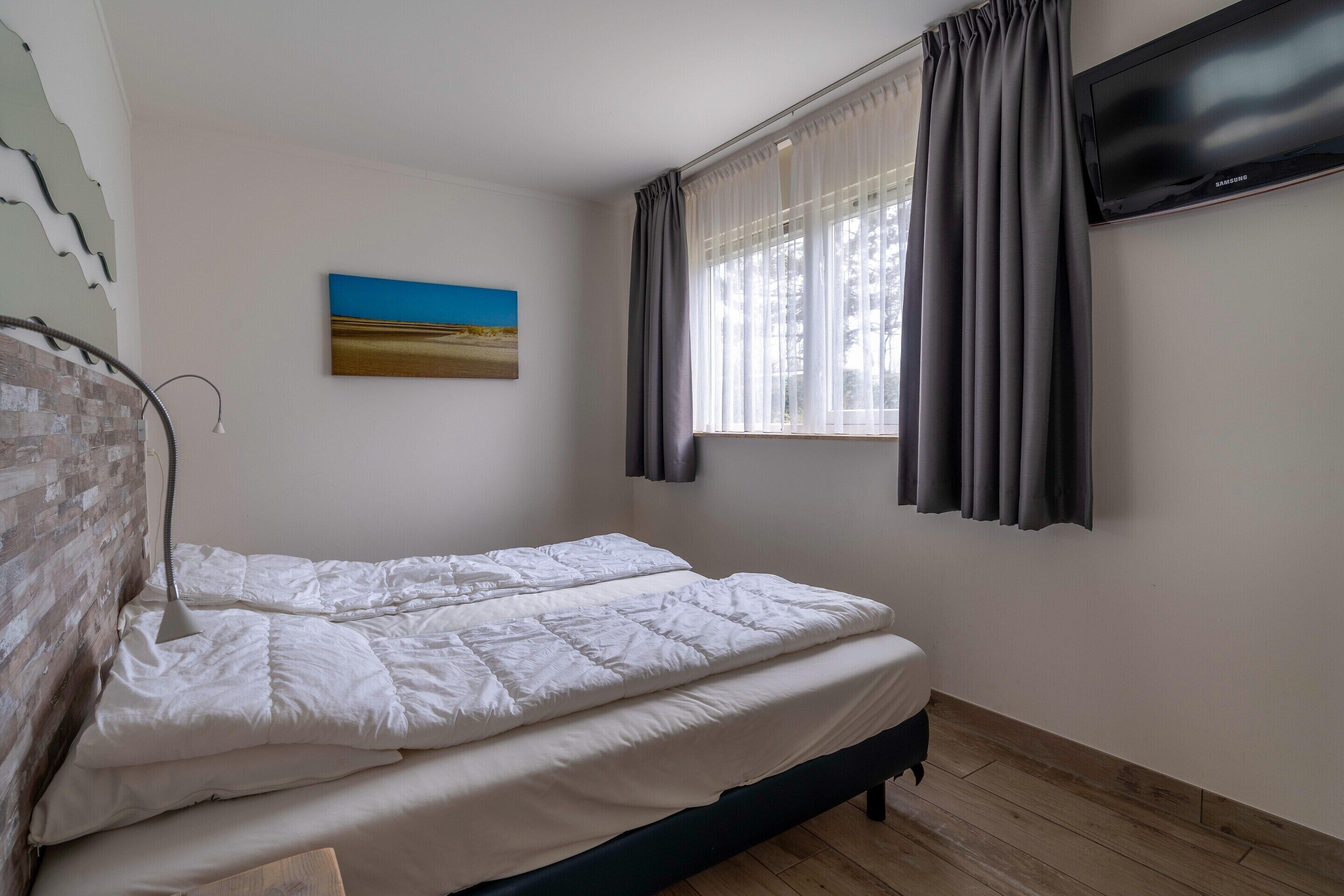 A bedroom with a bed, a window with curtains, a flat-screen TV, and a painting on the wall.