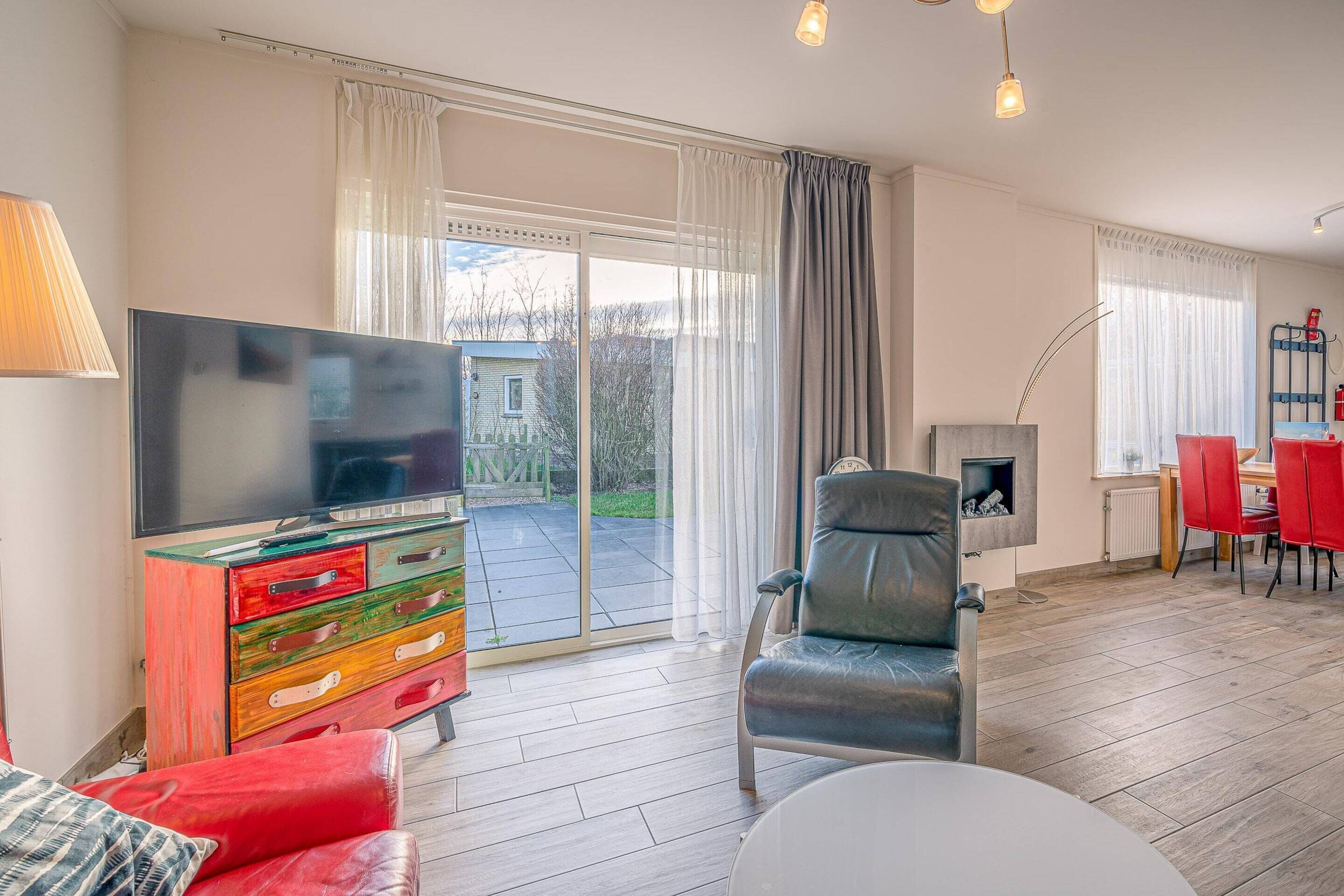 A modern living room with a flat-screen TV, a fireplace, a dining area with red chairs, and a large sliding glass door leading to a patio.