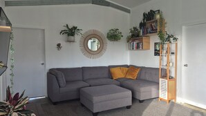A modern living room with a grey sectional sofa, a wooden ladder bookshelf, and various potted plants. - Spacious Valley Views Near City & Coast (Whareora)