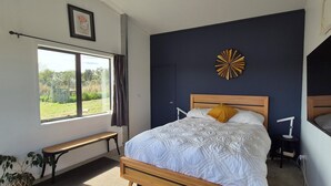 A bedroom with a large bed, a bench, a window with a view, and a wall clock. - Spacious Valley Views Near City & Coast (Whareora)