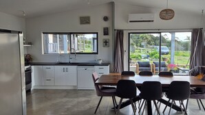 A modern kitchen with white cabinets, a dining area with a wooden table and black chairs, and large windows offering a view of the outdoors. - Spacious Valley Views Near City & Coast (Whareora)