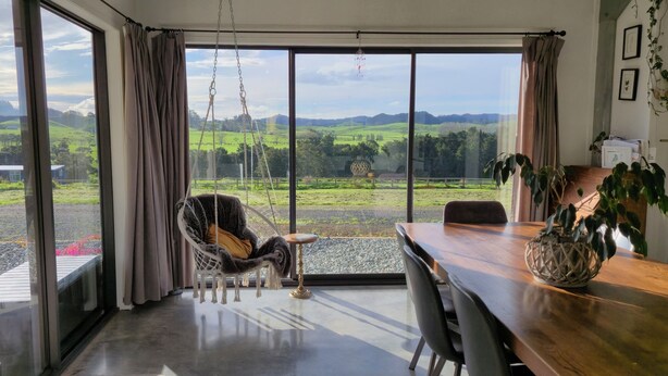 A room with a large sliding glass door leading to a view of green fields and distant hills. Inside, there's a wooden dining table with chairs, a hanging chair with a cushion, and a potted plant on the table. - Spacious Valley Views Near City & Coast (Whareora)
