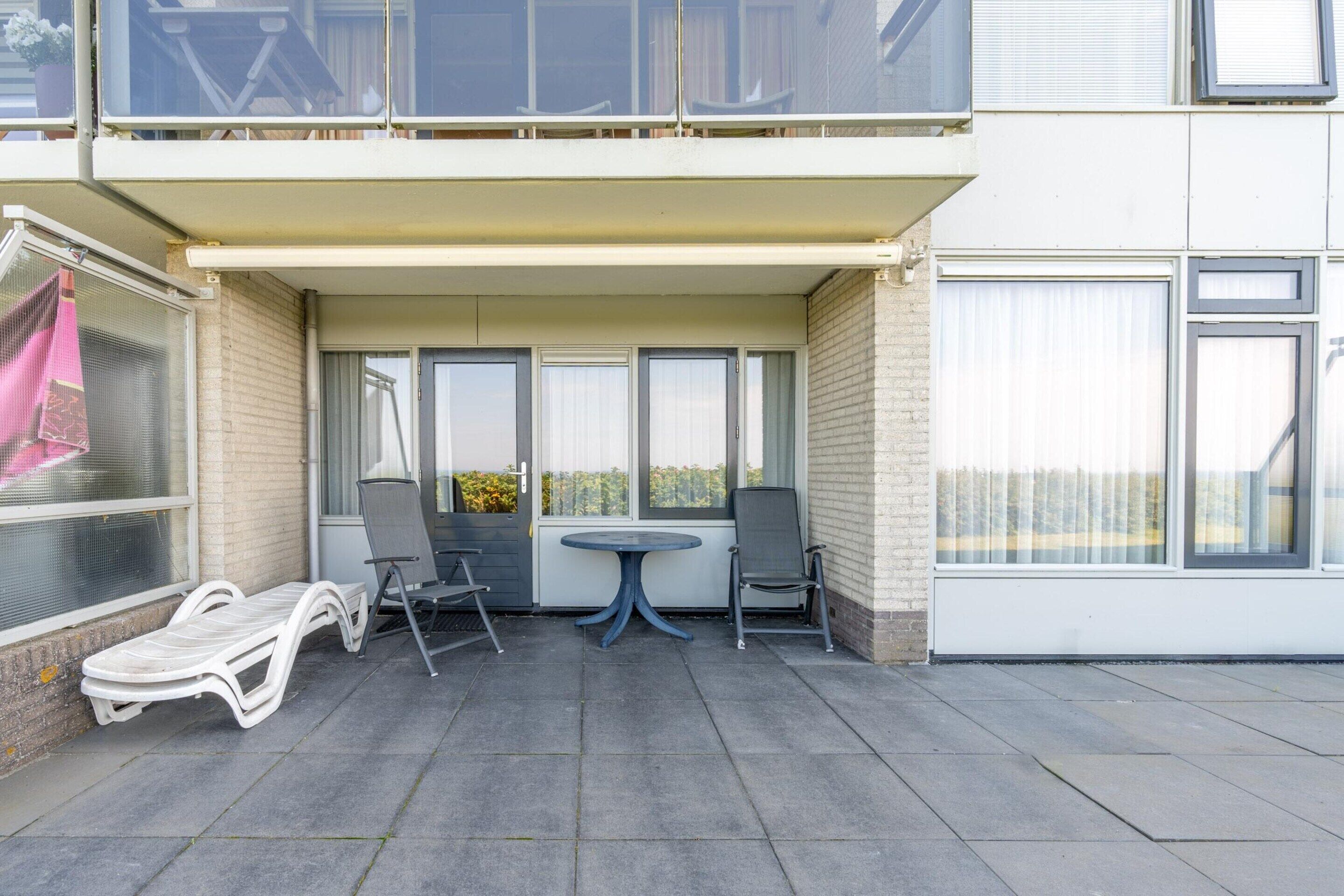 An outdoor balcony with a table and chairs, a sun lounger, and a view of a building in the background.
