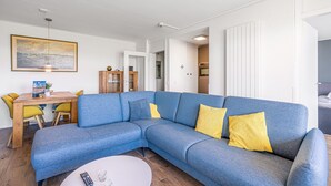 Een moderne woonkamer met een blauwe modulaire bank, gele kussens, een houten eettafel en een witte salontafel.