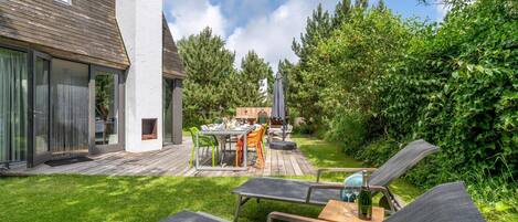 A modern outdoor patio with a dining table and chairs, a lounge area with two chairs, and a wooden deck.