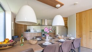 Un espace repas moderne avec une table en bois dressée pour quatre personnes, entourée de chaises. Un grand luminaire suspendu blanc est accroché au-dessus, et un vase rempli de fleurs est posé sur la table.