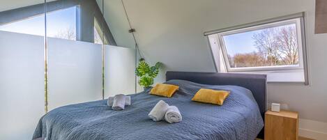 Quarto com uma cama grande, duas almofadas, uma mesinha de cabeceira, uma janela e uma planta.