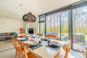 Uma sala de jantar com uma mesa grande posta para quatro pessoas, cercada por cadeiras de madeira. Uma luminária pendente moderna se destaca sobre a mesa.