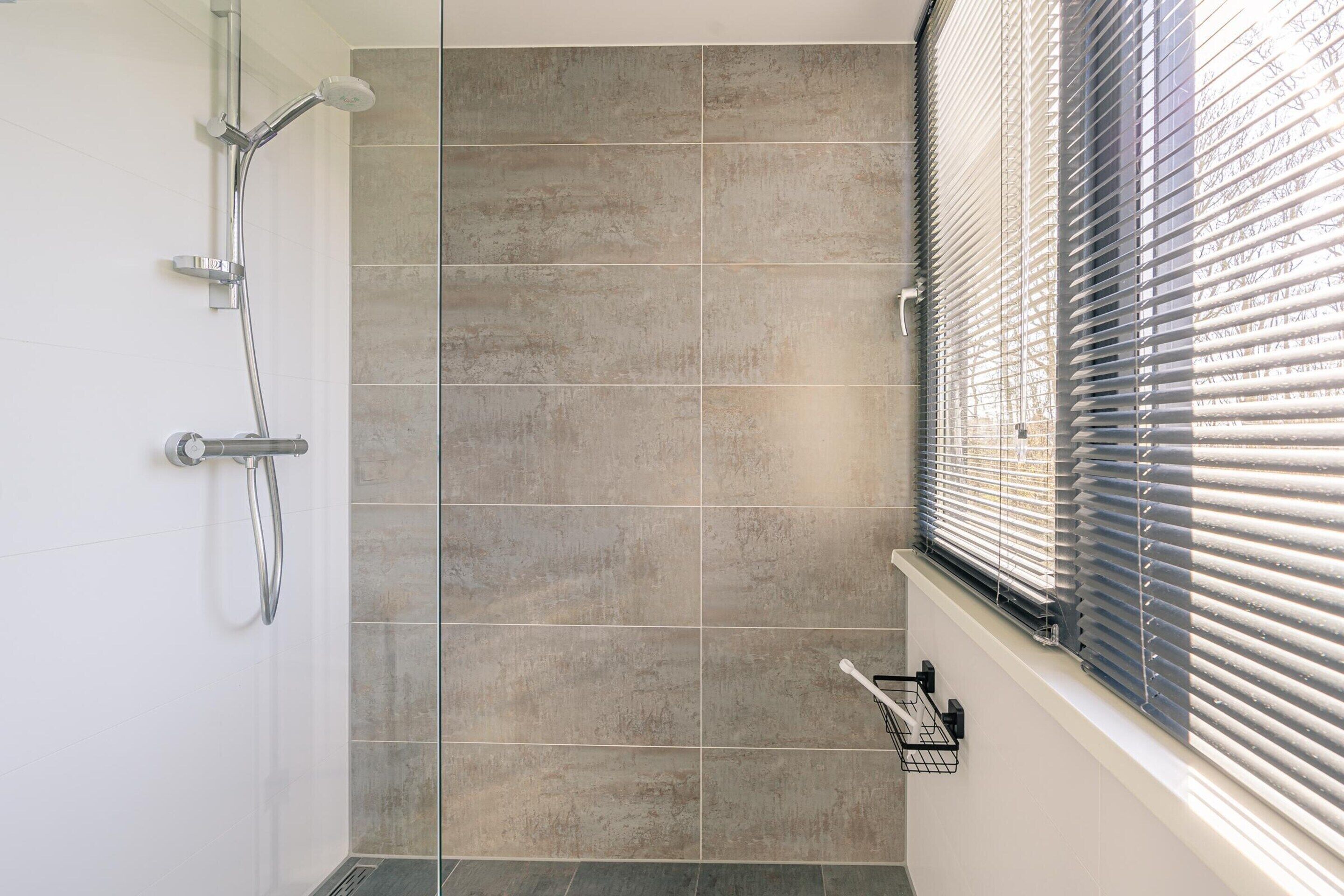 Une salle de bain avec une douche, des murs carrelés et une fenêtre avec des stores.