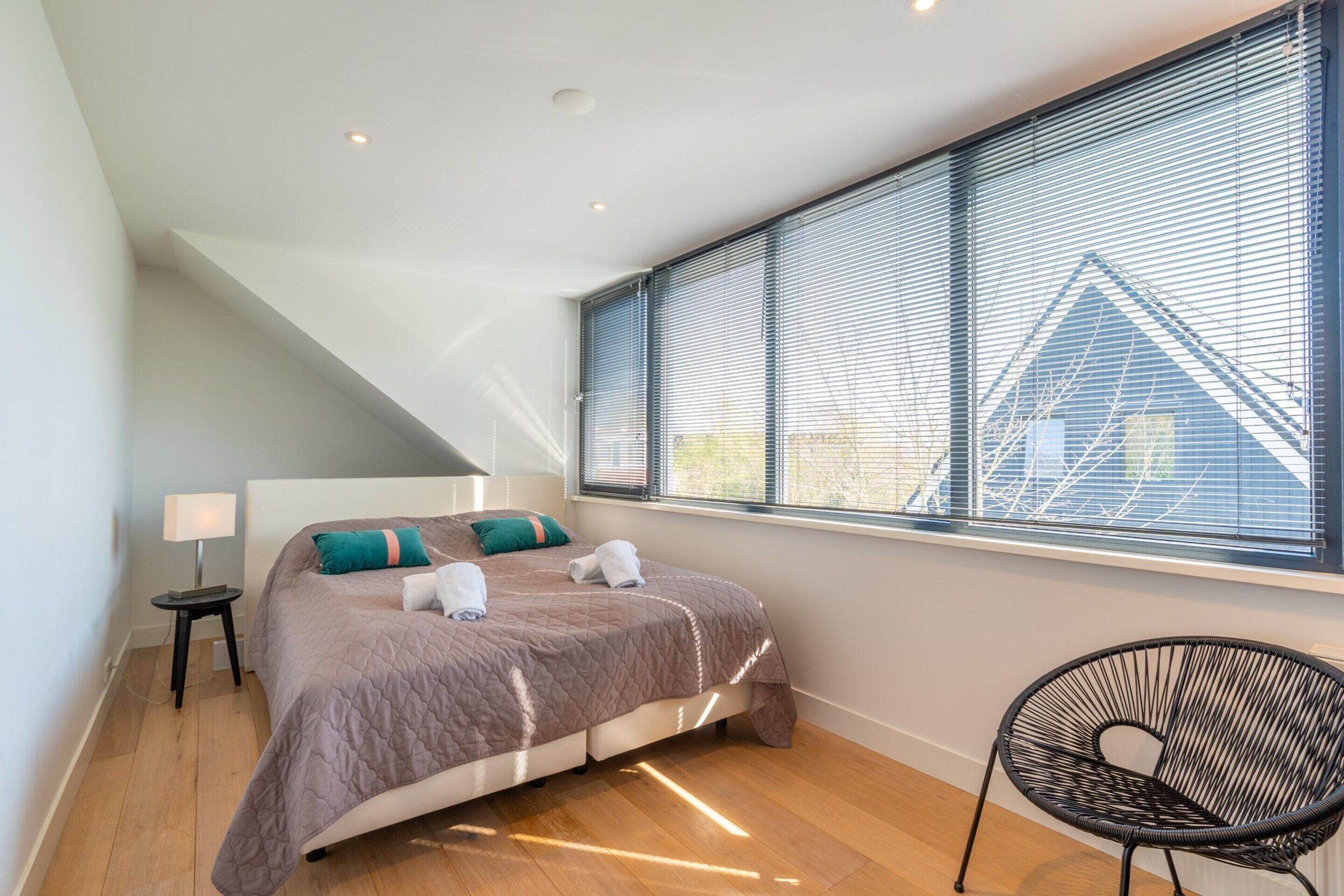 Ein Schlafzimmer mit Bett, Stuhl, Nachttisch und einem Fenster mit Jalousien.