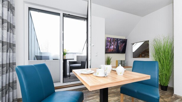 Een moderne kamer met een houten tafel, twee blauwe stoelen, een televisie aan de muur en een grote plant.