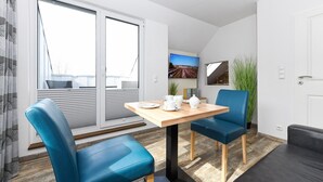 Een moderne kamer met een houten tafel, twee blauwe stoelen, een zwarte bank, een televisie en een groot raam met jaloezieën.