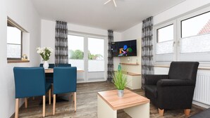 A modern living room with a wooden floor, a TV mounted on the wall, a dining area with blue chairs, a black armchair, and a small table with a plant. - Ferienwohnung in Bensersiel, 300 m vom Strand Entfernt (Esens)