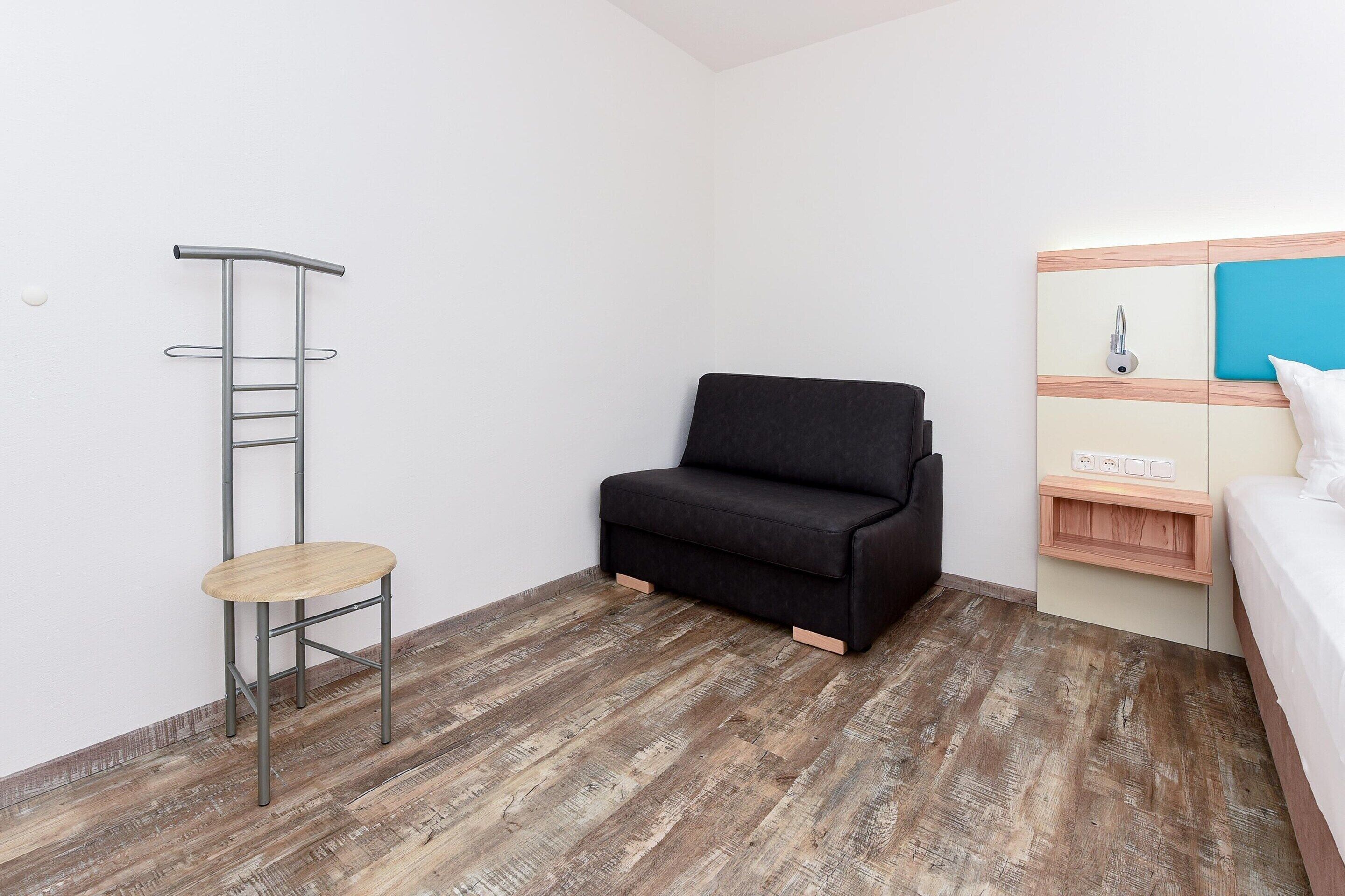 A small, modern room with a wooden floor, a black sofa, a small wooden chair, and a metal ladder against the wall.