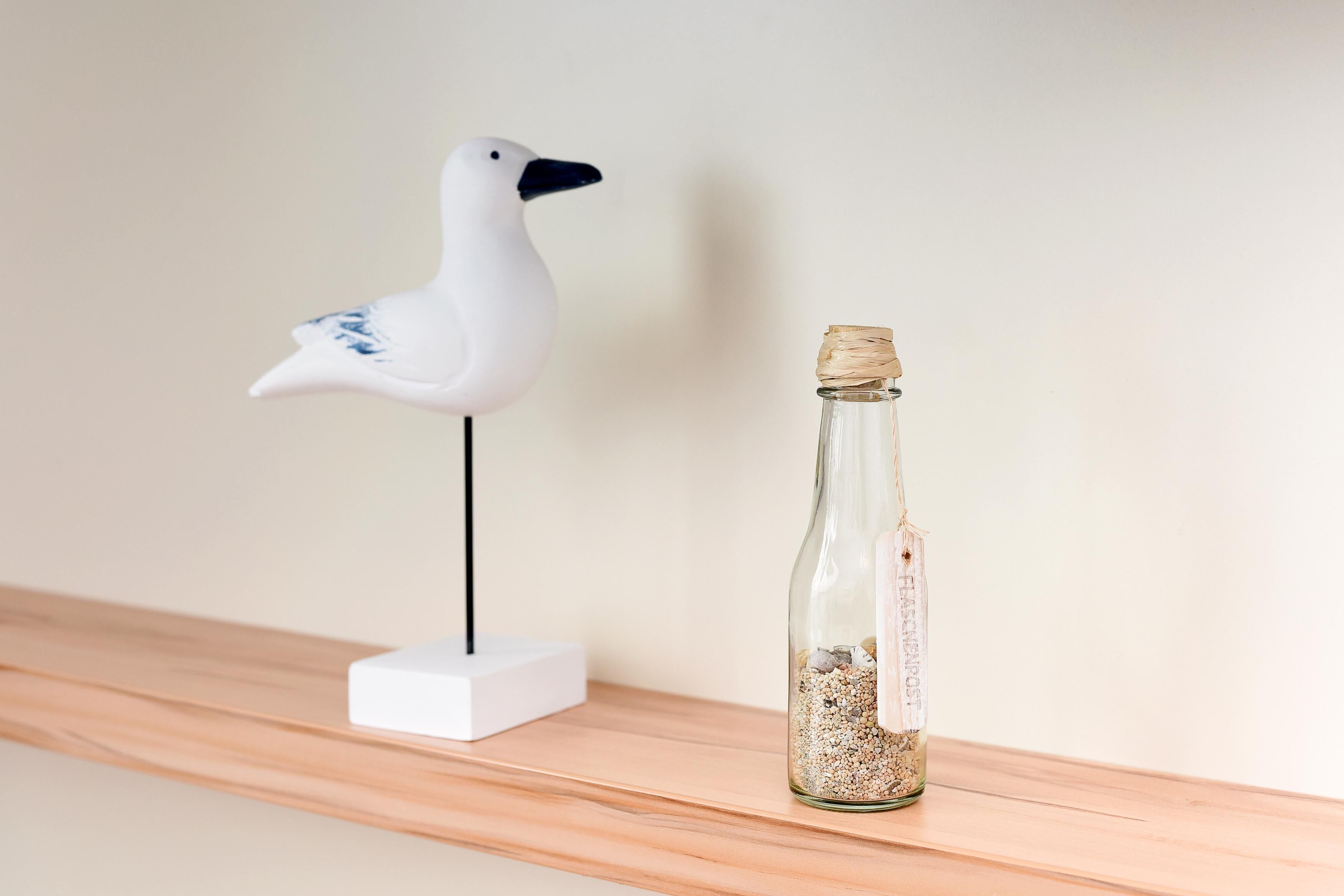 Een witte meeuwenbeeldje op een witte voet, naast een heldere glazen fles met kurk en zand.
