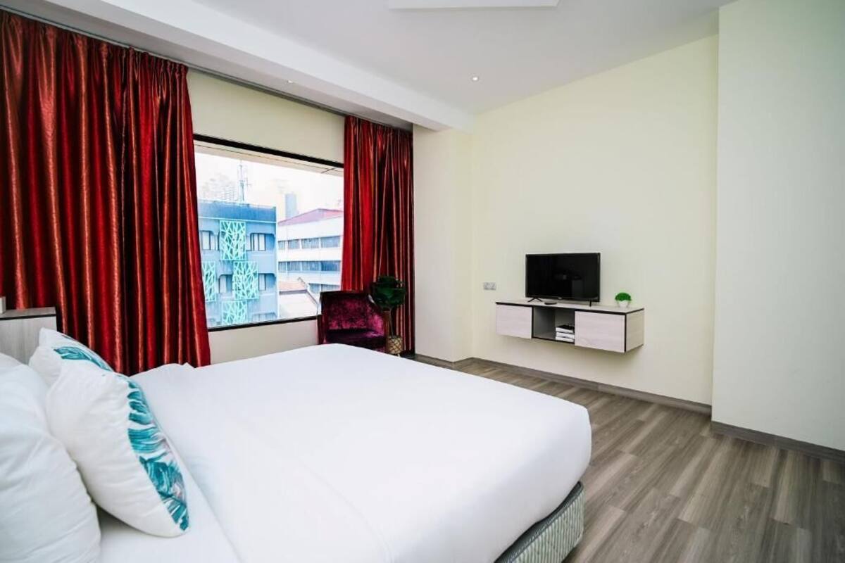 A hotel room with a large bed, red curtains, a TV on a white cabinet, and a view of buildings outside.