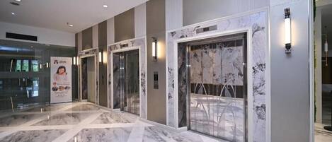 A modern hotel lobby with marble flooring, glass doors, and a glass elevator.