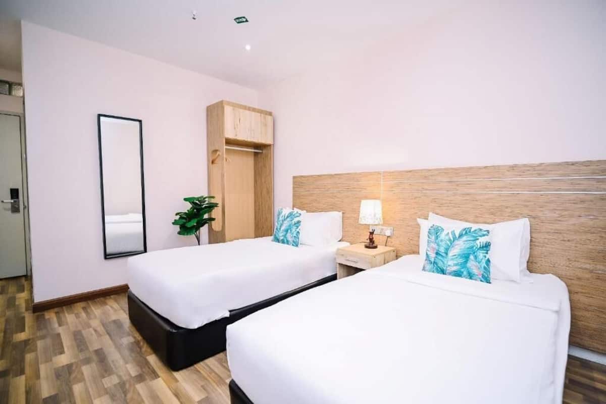 Habitación de hotel con dos camas, cabecero de madera, mesita de noche con lámpara, espejo y una planta.