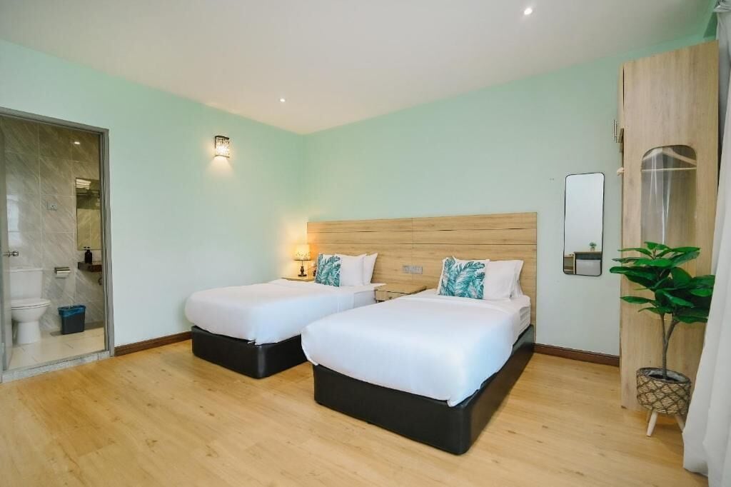 Hotelkamer met twee bedden, een houten hoofdbord, een kamerplant en een zichtbare badkamer door een glazen deur.