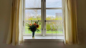 A room with a large window featuring a view of a garden and a building. There is a vase with flowers on the windowsill.