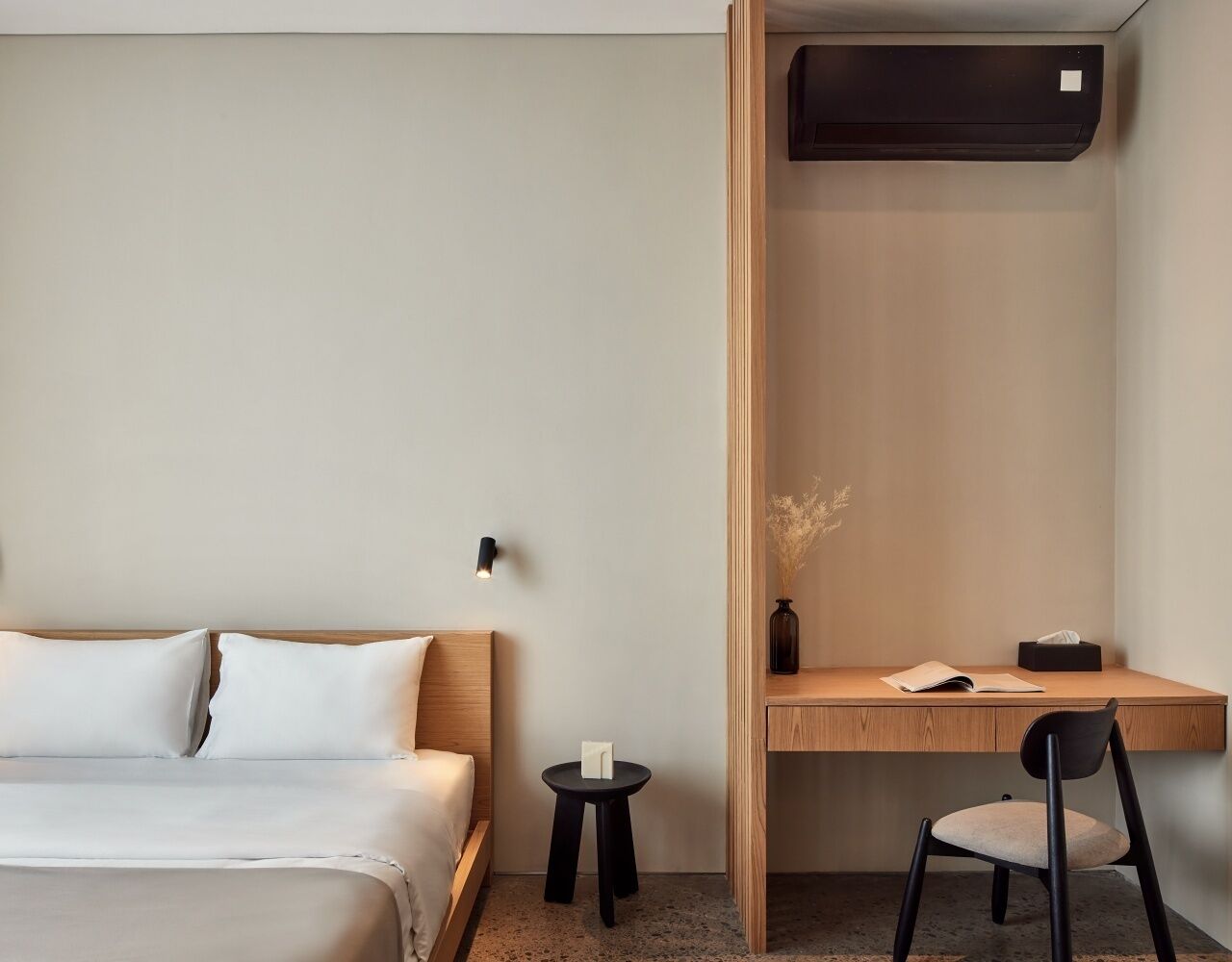 A hotel room with a bed, a desk with a chair, a small table, and a wall-mounted air conditioner.