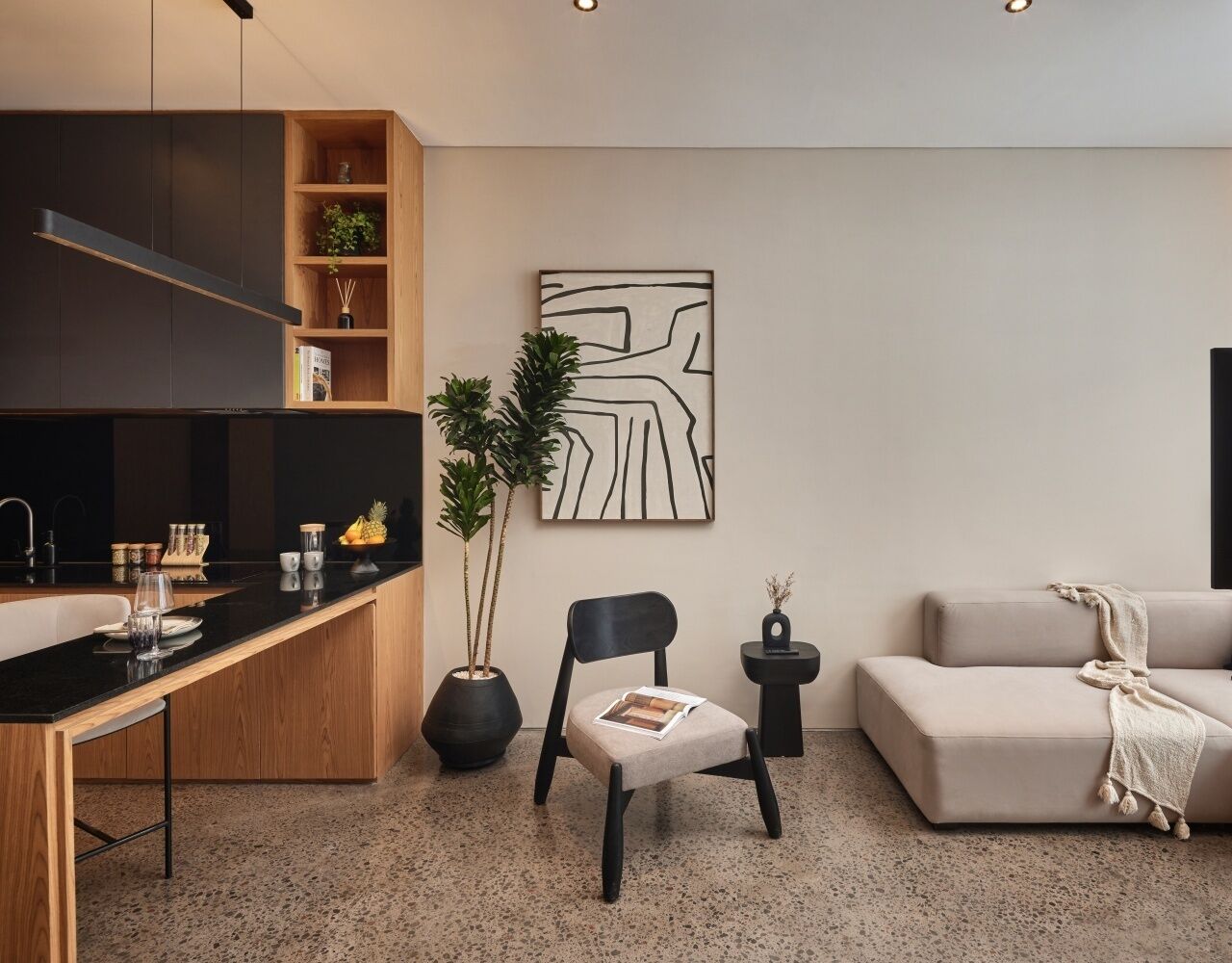 A modern living room with a kitchen area, featuring a black and wooden cabinet, a black chair with a magazine, a beige sofa, and a large abstract painting on the wall.