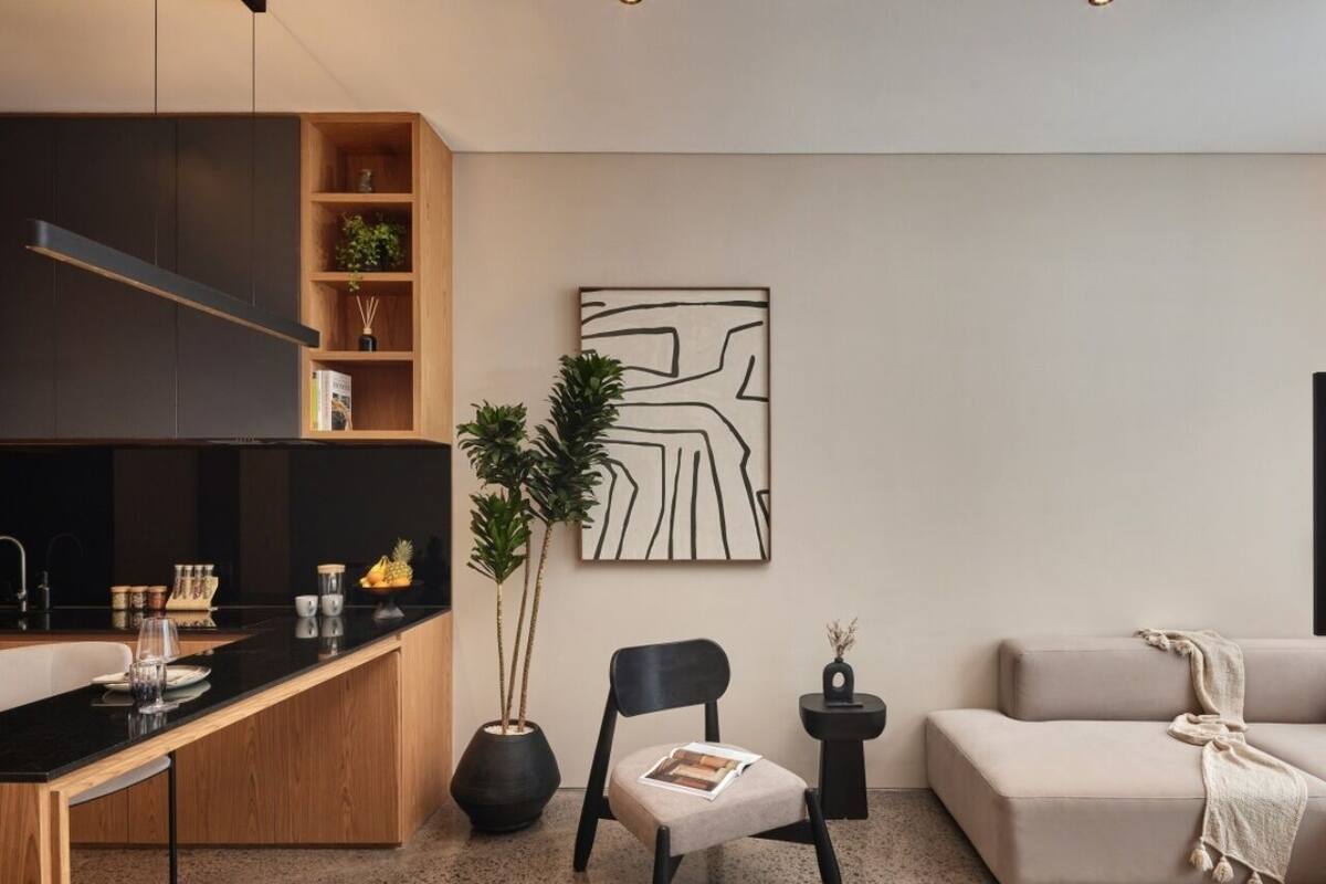 Ein modernes Wohnzimmer mit Küchenbereich, ausgestattet mit einem Holzkasten, einem schwarzen Sessel, einem beigen Sofa und einem minimalistischen Kunstwerk an der Wand.
