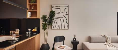 Ett modernt vardagsrum med ett köksområde, inrett med ett träskåp, en svart stol, en beige soffa och ett minimalistiskt konstverk på väggen.