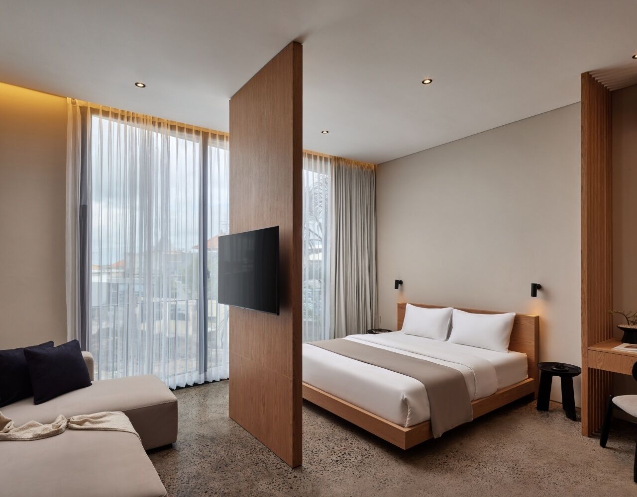 A modern hotel room with a large bed, a flat-screen TV mounted on a wooden panel, a sofa, and a desk with a chair.