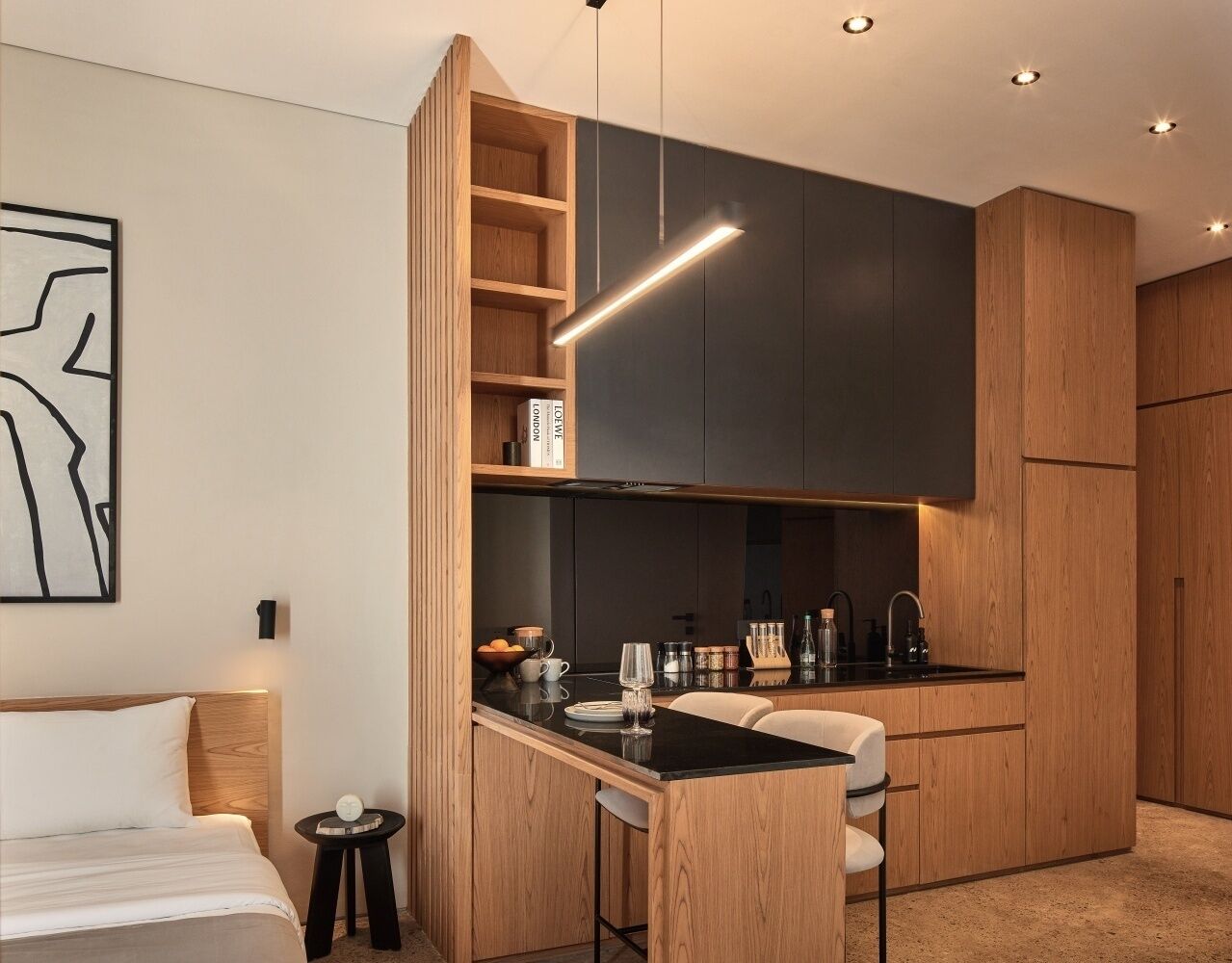 A modern hotel room with a kitchenette, a bed with white bedding, a small bedside table, and a wall-mounted artwork.