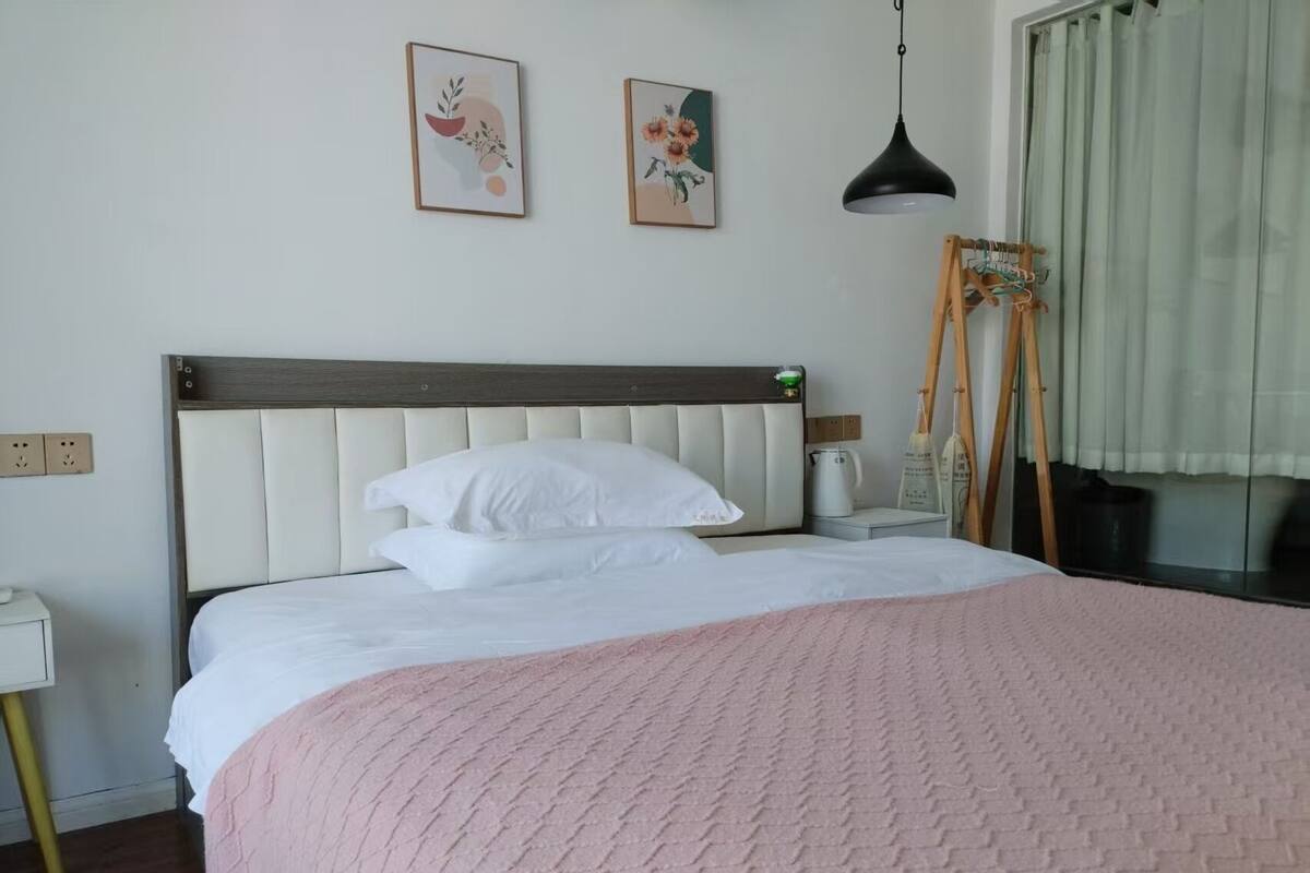 Una cama bien hecha con sábanas blancas y una manta rosa, una mesita de noche con lámpara, y dos cuadros enmarcados en la pared.