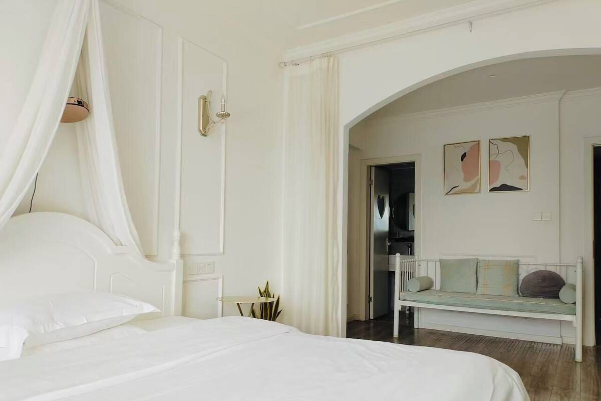 Habitación de hotel con una cama grande, una mesita de noche con lámpara, un banco con cojines y una puerta que da a otra habitación.