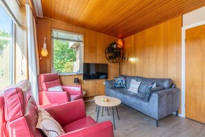 Una sala de estar acogedora con techo de madera, un sofá gris, sillones de cuero rojo, una mesita y un televisor montado en la pared.
