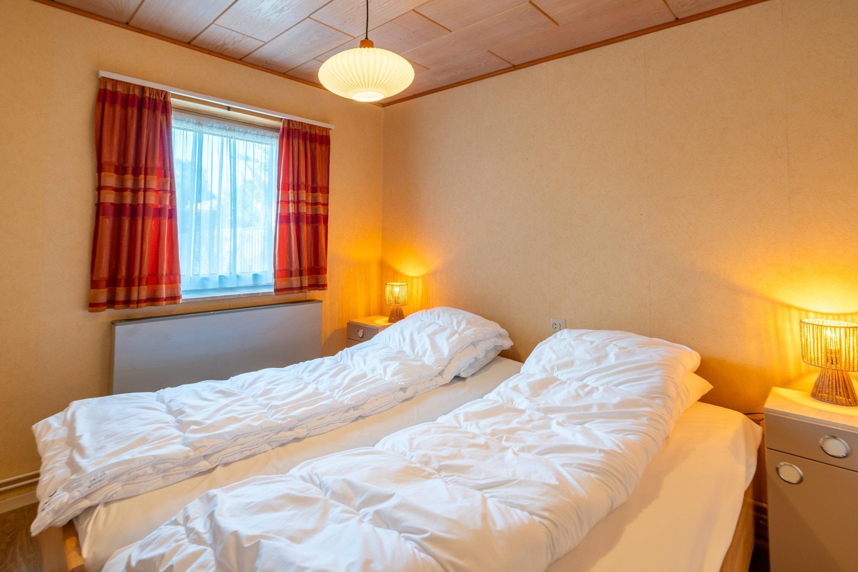 A bedroom with two single beds, a window with curtains, a bedside lamp, and a ceiling light fixture.