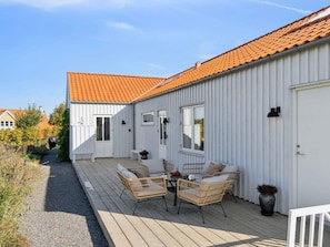 En hvid bygning med rødt tegltag, en overdækket terrasse med fletmøbler og en grussti, der fører til indgangen.