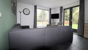 A living room with a grey sofa, a wall clock, a flat-screen TV, and a window with curtains. - Modern Bungalow in Ewijk for 6 (Ewijk)