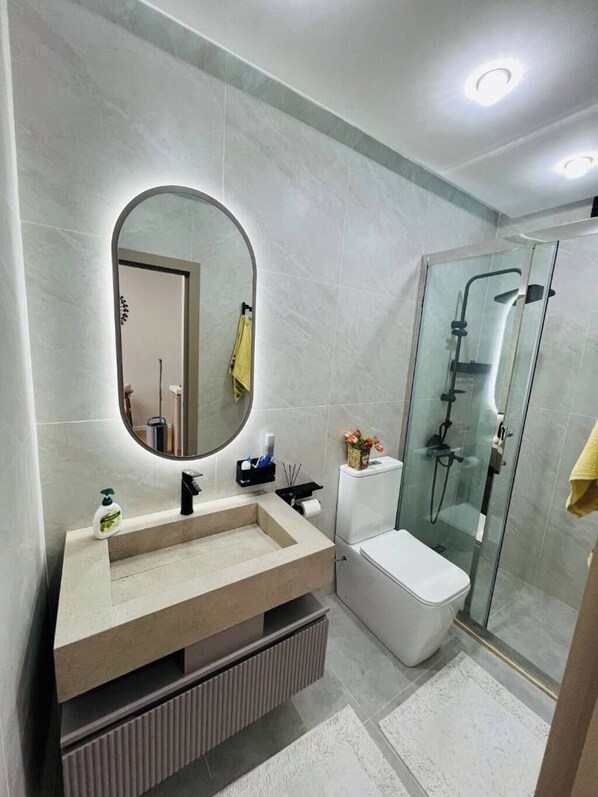 A modern bathroom with a large oval mirror, a beige vanity, a white toilet, and a glass-enclosed shower. - Lev iki (Baku)