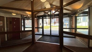 Une entrée dotée d’un cadre en bois et de portes vitrées, qui donne sur un espace extérieur couvert offrant une vue sur les bâtiments et un parking.