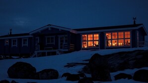 Casa de dos pisos con amplios ventanales en la planta baja, rodeada de tierra y rocas cubiertas de nieve.