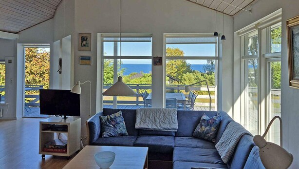 A living room with a sofa, a coffee table, and a television. There are large windows with a view of the outdoors.
