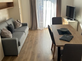 Ein modernes Wohnzimmer mit grauem Sofa, Esstisch aus Holz und einem Fernseher auf einem Schrank.