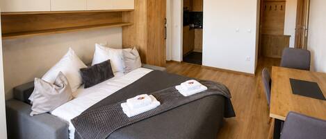 A modern hotel room with a bed, a sofa, a dining table, and a kitchenette.