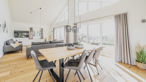 Un salon spacieux avec une table à manger en bois, des canapés gris et une grande fenêtre donnant sur l’extérieur.