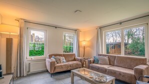Een ruime woonkamer met twee beige banken, een glazen salontafel en een vloerlamp. Grote ramen bieden uitzicht op groen buiten.