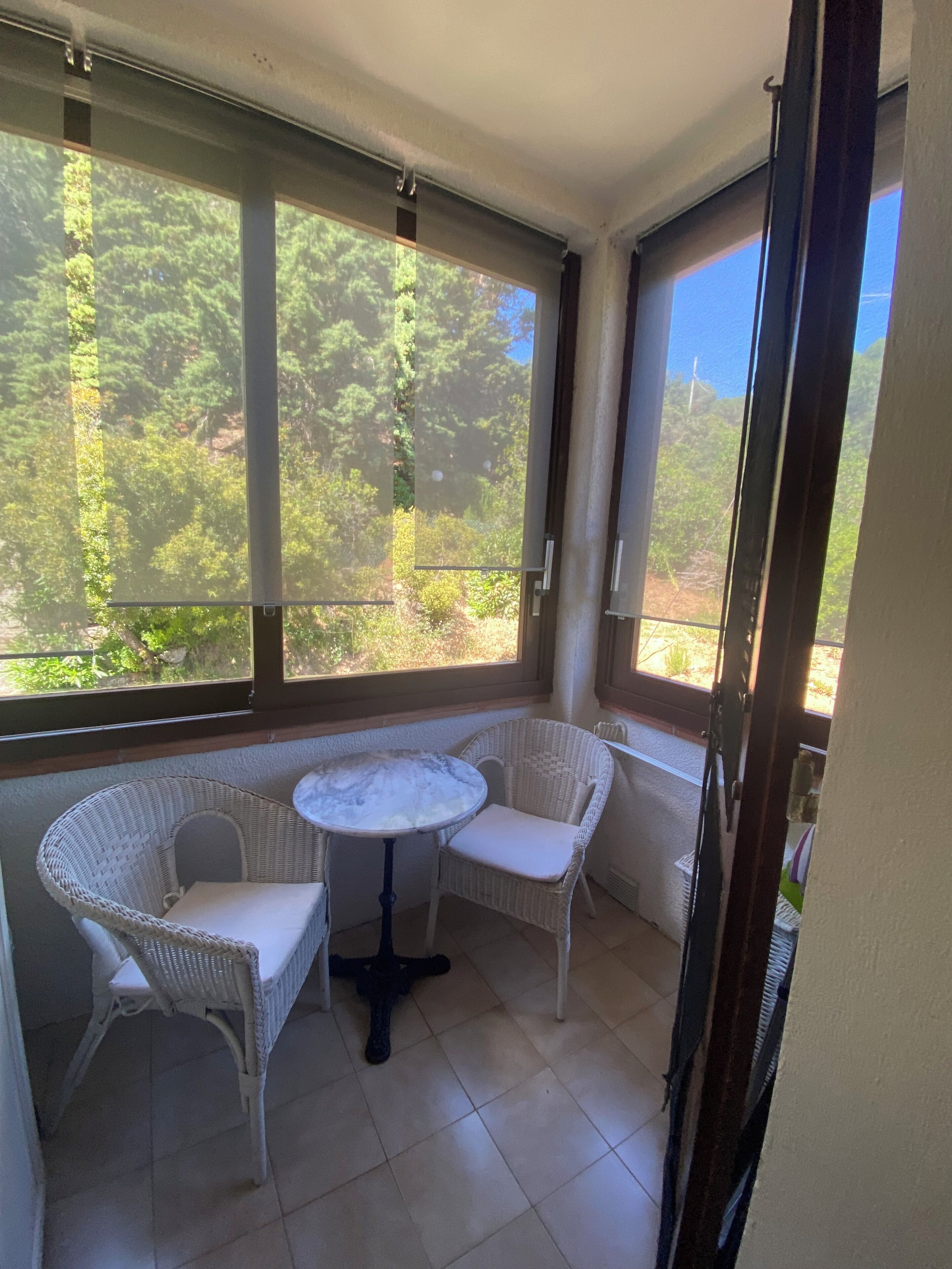 Una habitación luminosa con una mesa y dos sillas de mimbre, ventanas amplias con vistas a la vegetación y suelo de baldosas.
