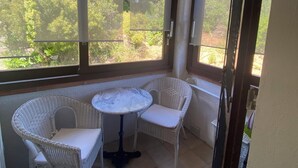 A sunlit room with a table and two wicker chairs, large windows with a view of greenery, and a tiled floor.